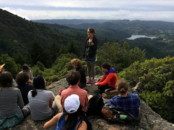 Workshop participants sit, while a facilitator stands on a rock outcropping with trees and vegetation, a lake and cloudy skies in the distance.