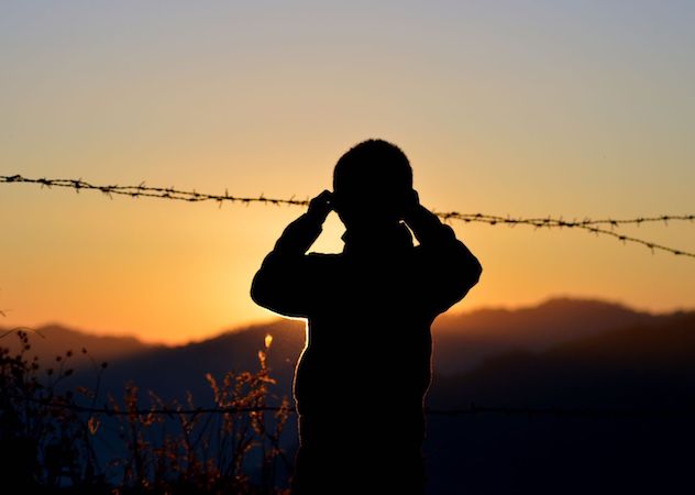 A child gazes through barbed wire into the distance of open, black hills and orange sunset.