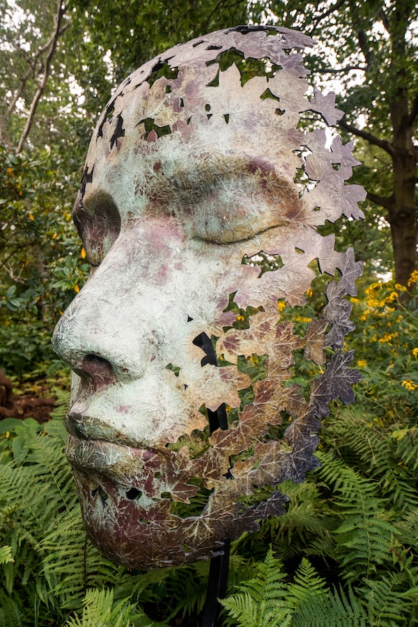 A sculpture of a face with eyes closed is situated in a forested setting.