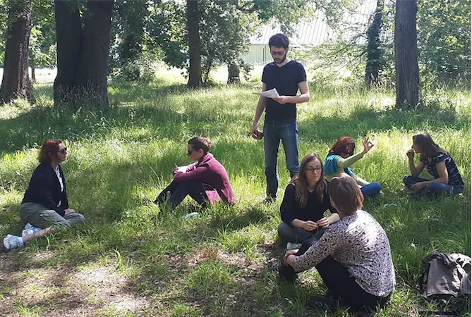 Humans gathered, sitting and standing, in grass in a forested area.