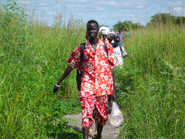 A line of humans with bags walk through a grassy, marshy area.